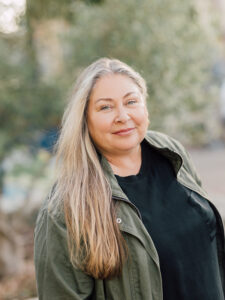 SLOMA Executive Director Leann Standish. A woman with long blonde hair wearing a green shirt and black jacket.