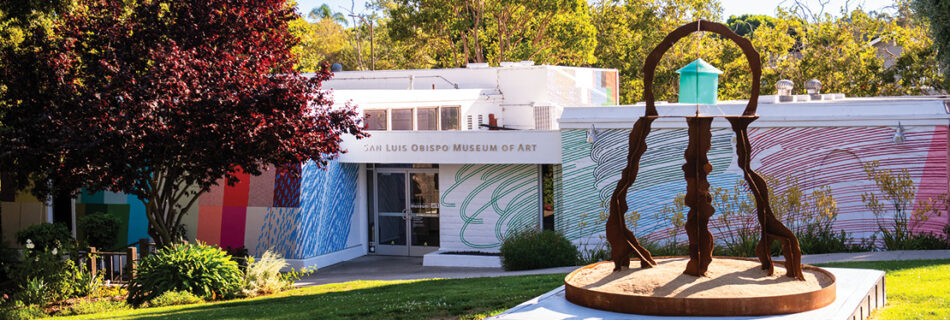 A steel sculpture reminiscent of a jellyfish with a teal resin "house" in the middle. SLOMA in the background with a colorful mural