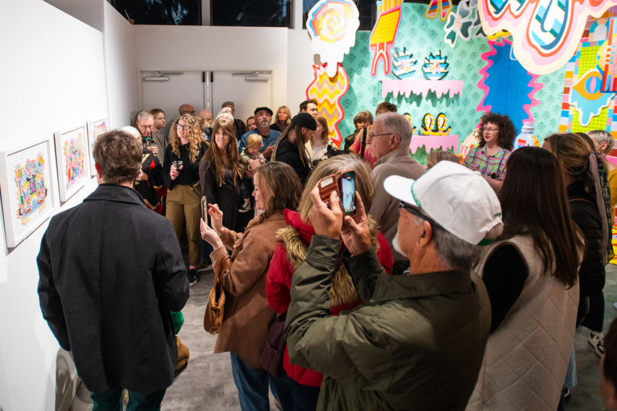 A large crowd fills a gallery with an art installation featuring intensely bright colors and patterns from floor to ceiling